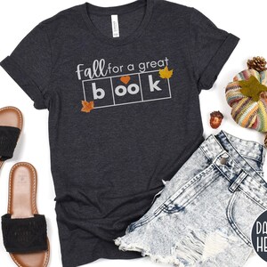 May include: Dark grey heather t-shirt with the text "Fall for a great book" in white letters. The text is in a box with a heart in the second box. There are small orange and yellow leaf graphics on the shirt.