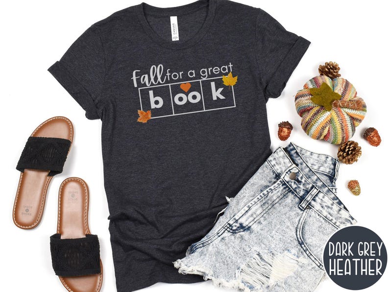 May include: Dark grey heather t-shirt with the text "Fall for a great book" in white letters. The text is in a box with a heart in the second box. There are small orange and yellow leaf graphics on the shirt.