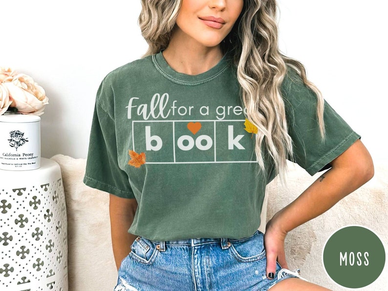 Puede incluir: Una camiseta verde con el texto "fall for a great book" en letras blancas. Las letras est&aacute;n en una caja con un coraz&oacute;n en la "o". Hay peque&ntilde;as hojas de oto&ntilde;o en las esquinas de la caja.