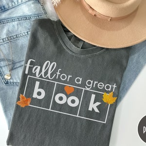 Puede incluir: Una camiseta gris con el texto "Fall for a great book" en letras blancas. La palabra "book" est&aacute; en cuadros individuales con un peque&ntilde;o coraz&oacute;n rojo en la "o". Hay peque&ntilde;as hojas naranjas a cada lado del texto.