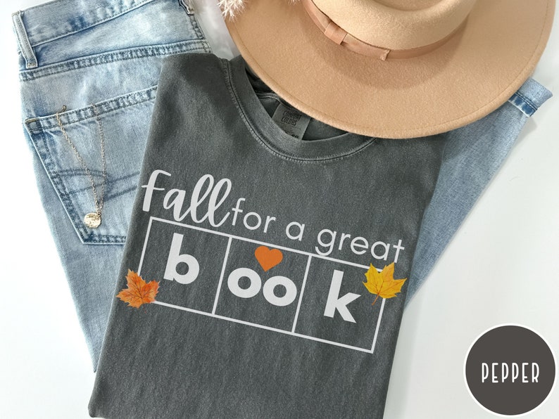 Puede incluir: Una camiseta gris con el texto "Fall for a great book" en letras blancas. La palabra "book" est&aacute; en cuadros individuales con un peque&ntilde;o coraz&oacute;n rojo en la "o". Hay peque&ntilde;as hojas naranjas a cada lado del texto.