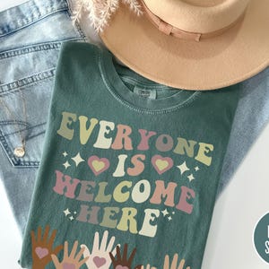 May include: A green t-shirt with the text "Everyone is welcome here" in a retro font with colourful hearts and stars. The shirt has a distressed look and features a design of hands with hearts in different skin tones.