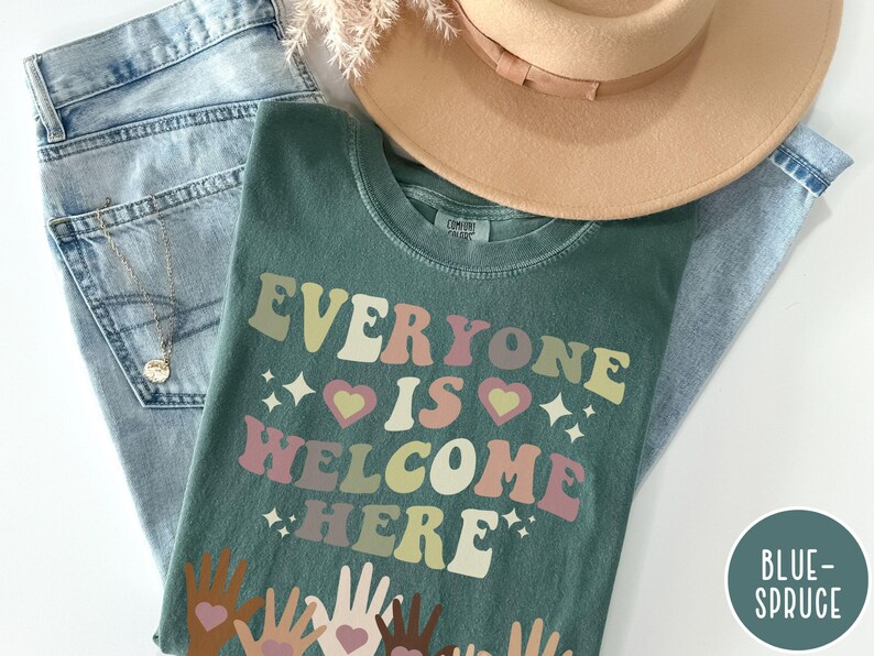 May include: A green t-shirt with the text "Everyone is welcome here" in a retro font with colourful hearts and stars. The shirt has a distressed look and features a design of hands with hearts in different skin tones.