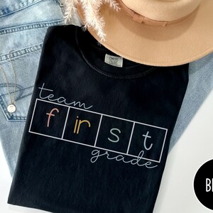 Op de afbeelding: Zwart T-shirt met de tekst "team first grade" in kleurrijke letters. De letters bevinden zich in afzonderlijke vakjes met een witte rand.