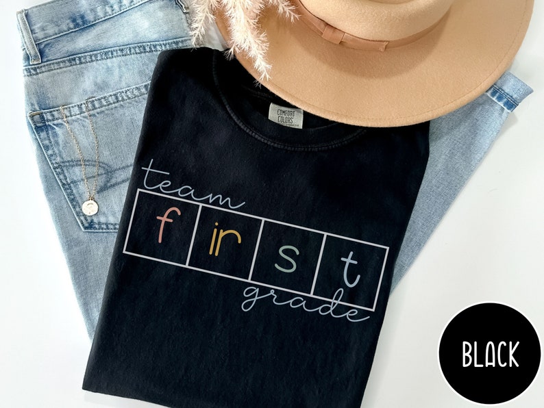 Op de afbeelding: Zwart T-shirt met de tekst "team first grade" in kleurrijke letters. De letters bevinden zich in afzonderlijke vakjes met een witte rand.