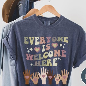 May include: A dark blue t-shirt with the text "Everyone is Welcome Here" in colourful retro lettering. The shirt features a design of five hands with hearts in the palms, symbolising inclusivity.
