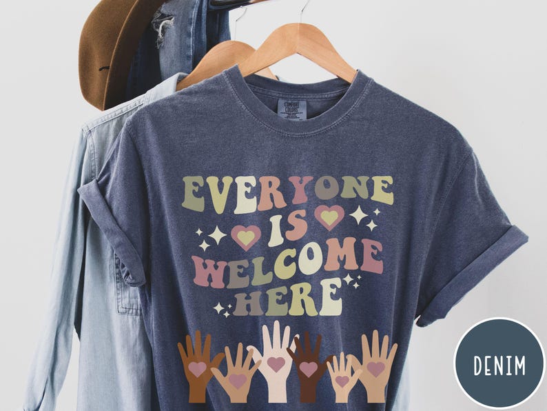 May include: A dark blue t-shirt with the text "Everyone is Welcome Here" in colourful retro lettering. The shirt features a design of five hands with hearts in the palms, symbolising inclusivity.