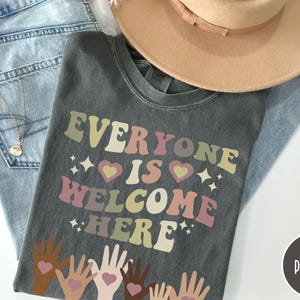 May include: A dark grey t-shirt with the text "Everyone is Welcome Here" in a retro font. The text is in shades of pink, yellow, and brown. There are small hearts and stars around the text. The bottom of the shirt features a design of hands of different skin tones reaching up with hearts in their palms.