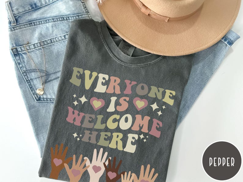 May include: A dark grey t-shirt with the text "Everyone is Welcome Here" in a retro font. The text is in shades of pink, yellow, and brown. There are small hearts and stars around the text. The bottom of the shirt features a design of hands of different skin tones reaching up with hearts in their palms.