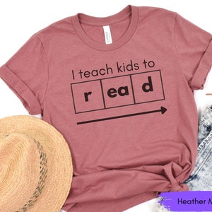 Może przedstawiać: Koszulka w kolorze heather mauve z napisem "I teach kids to read" w czarnych literach. Tekst jest wyświetlany w stylu układu okresowego z literami "r", "ea" i "d" w oddzielnych polach.
