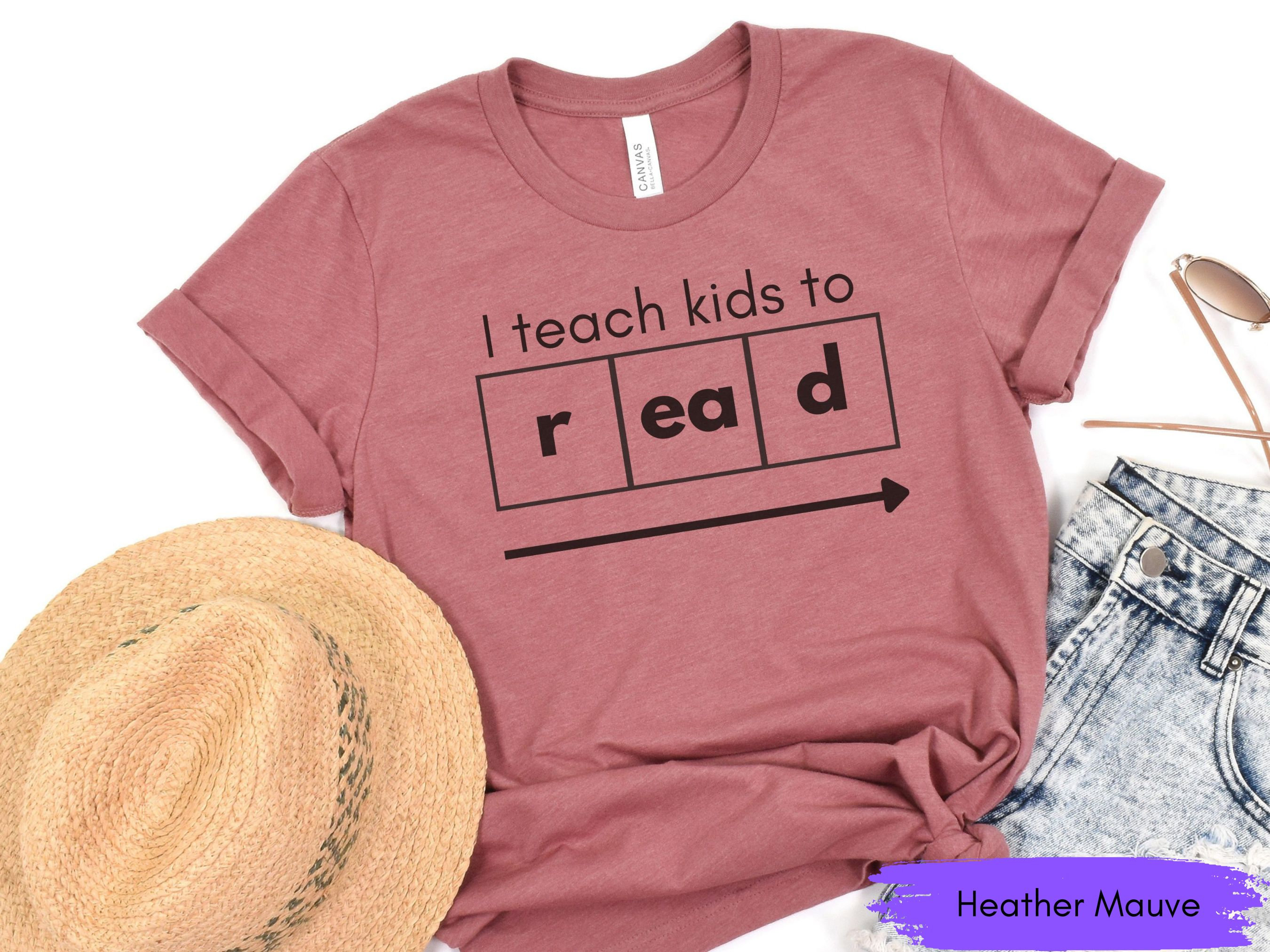 Może przedstawiać: Koszulka w kolorze heather mauve z napisem "I teach kids to read" w czarnych literach. Tekst jest wyświetlany w stylu układu okresowego z literami "r", "ea" i "d" w oddzielnych polach.