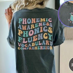 May include: A dark gray t-shirt with the words "phonemic awareness, phonics, fluency, vocabulary, comprehension" printed in rainbow colors on the back. The front of the shirt has a small graphic of a book with the text "Reading is a right" printed around it.