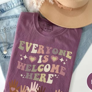 May include: A purple t-shirt with the text "Everyone is Welcome Here" in a retro font. The text is in shades of pink, yellow, and green. There are small hearts and stars around the text. The shirt is lying on a pair of blue jeans and a brown hat.