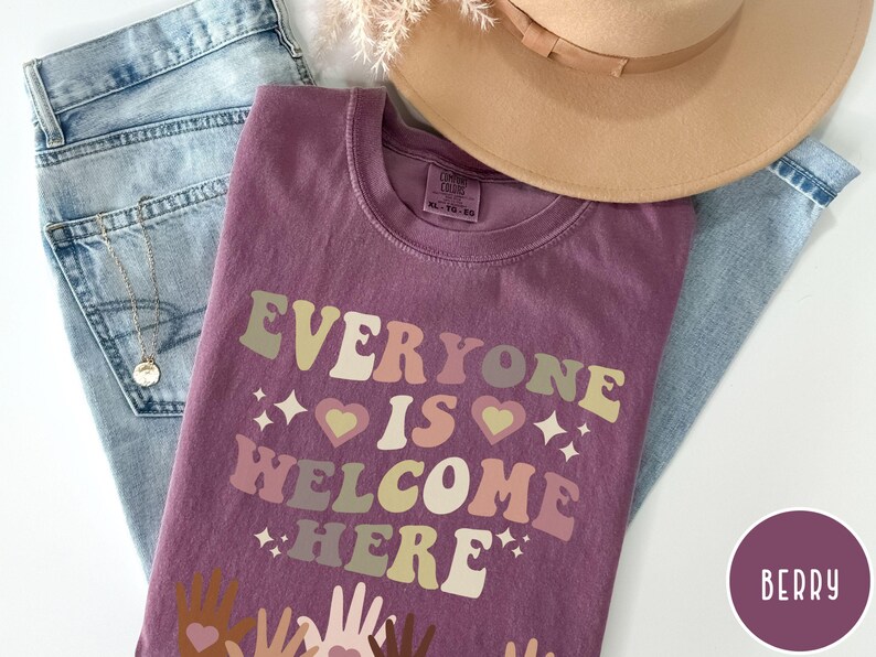 May include: A purple t-shirt with the text "Everyone is Welcome Here" in a retro font. The text is in shades of pink, yellow, and green. There are small hearts and stars around the text. The shirt is lying on a pair of blue jeans and a brown hat.
