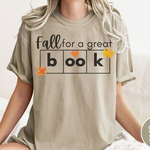 Puede incluir: Una camiseta beige con el texto "Fall for a great book" en letras negras. El texto est&aacute; en un cuadro con un coraz&oacute;n y hojas de oto&ntilde;o.