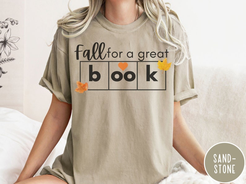 Puede incluir: Una camiseta beige con el texto "Fall for a great book" en letras negras. El texto est&aacute; en un cuadro con un coraz&oacute;n y hojas de oto&ntilde;o.