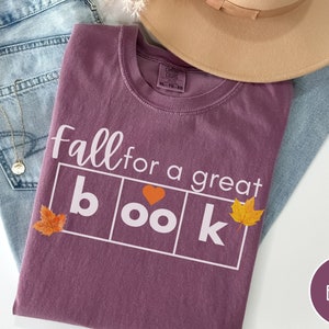 Puede incluir: Una camiseta color granate con el texto "Fall for a great book" en blanco. La palabra "book" est&aacute; en cuadros blancos con un coraz&oacute;n y hojas de oto&ntilde;o en cada cuadro.