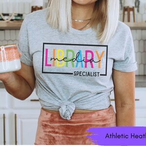 May include: A gray t-shirt with the text "LIBRARY MEDIA SPECIALIST" in a colorful rainbow font. The shirt is tied at the waist and the model is wearing a pink skirt. The text "Athletic Heather" is visible at the bottom of the image.