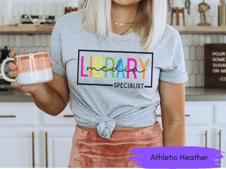 May include: A gray t-shirt with the text "LIBRARY MEDIA SPECIALIST" in a colorful rainbow font. The shirt is tied at the waist and the model is wearing a pink skirt. The text "Athletic Heather" is visible at the bottom of the image.