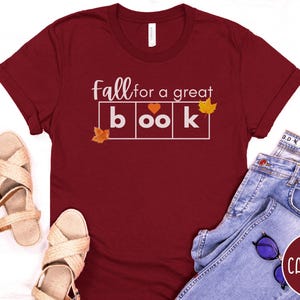 May include: A maroon t-shirt with the text "Fall for a great book" in white letters. The text is in a box with a heart in the middle of the word "book". There are small orange leaf graphics on either side of the text.