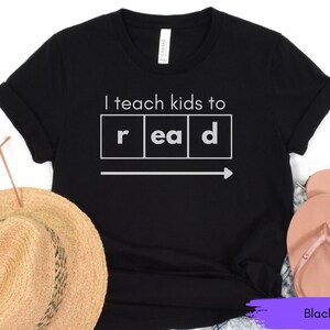 Może przedstawiać: Czarny T-shirt z napisem "Uczę dzieci czytać" w białych literach. Słowo "czytać" jest podzielone na poszczeg&oacute;lne litery w ramkach.