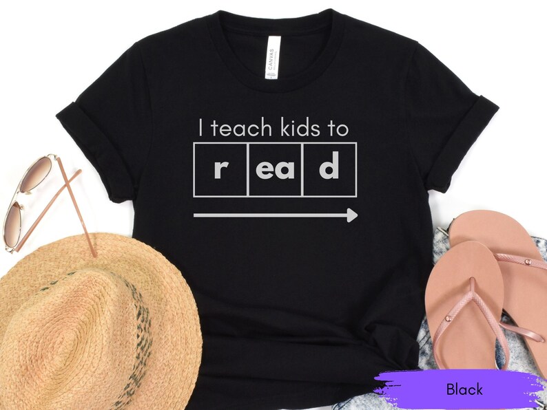 Może przedstawiać: Czarny T-shirt z napisem "Uczę dzieci czytać" w białych literach. Słowo "czytać" jest podzielone na poszczeg&oacute;lne litery w ramkach.