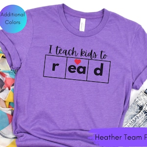 May include: Purple t-shirt with the text "I teach kids to read" with a heart in the word "read". The shirt is heather team purple.