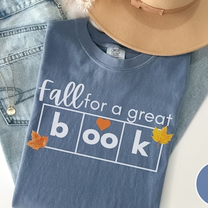 Puede incluir: Una camiseta azul vaquero con el texto "fall for a great book" en blanco. La palabra "book" est&aacute; en un cuadro con un coraz&oacute;n y hojas de oto&ntilde;o a cada lado.
