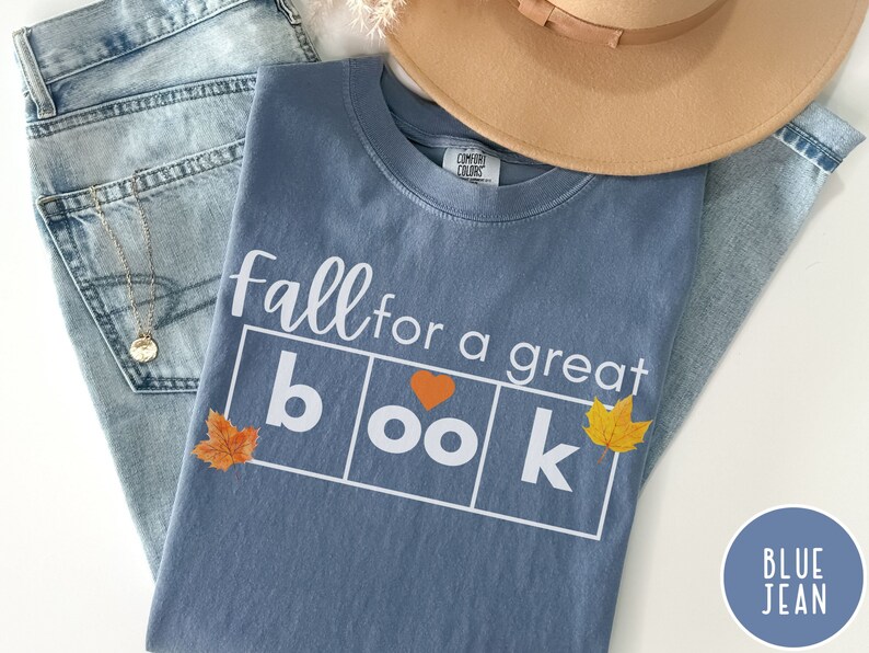 Puede incluir: Una camiseta azul vaquero con el texto "fall for a great book" en blanco. La palabra "book" est&aacute; en un cuadro con un coraz&oacute;n y hojas de oto&ntilde;o a cada lado.