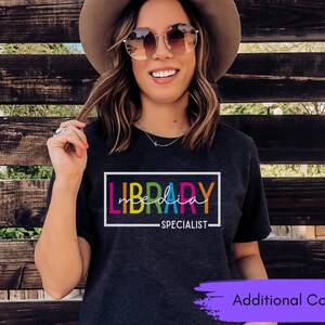 May include: A dark grey t-shirt with the text "LIBRARY SPECIALIST" in a rainbow color scheme. The text is inside a white rectangle.