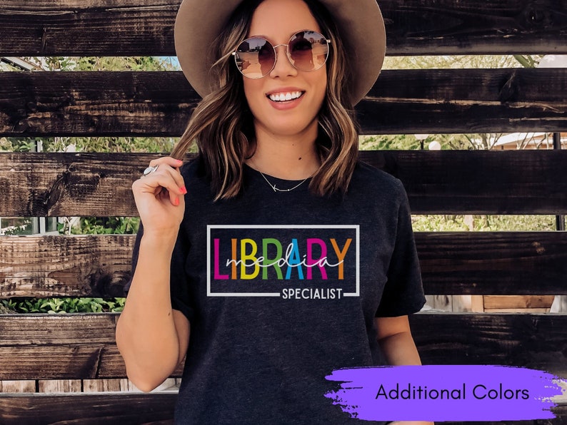 May include: A dark grey t-shirt with the text "LIBRARY SPECIALIST" in a rainbow color scheme. The text is inside a white rectangle.