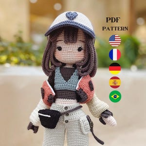 Puede incluir: Muñeca de crochet con cabello castaño que lleva un sombrero blanco y azul, una chaqueta marrón y naranja, un top gris, pantalones blancos y botas marrones. La muñeca lleva una bolsa negra. La imagen incluye el texto "PDF PATTERN" y banderas de Estados Unidos, Francia, Alemania, España y Brasil.