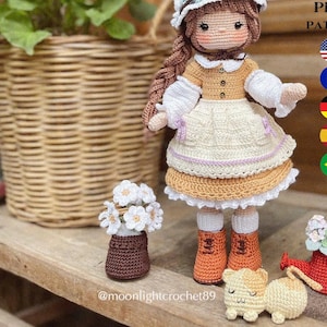 以下が含まれることがあります： かぎ針編み人形のパターンPDF。人形は緑色の帽子をかぶった茶色と白のドレスを着ています。人形は白い花のブーケを持っています。パターンには、アメリカ、フランス、ドイツ、スペイン、ブラジルの国旗が含まれています。