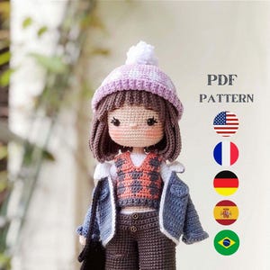 以下が含まれることがあります： 英語、フランス語、ドイツ語、スペイン語、ポルトガル語で書かれた、かぎ針編み人形のパターンPDF。人形は、紫と白の帽子、チェック柄のシャツ、青いデニムジャケット、茶色のズボン、黒い靴を履いています。人形は黒いバッグを持っています。