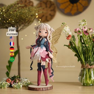 Peut inclure: Une poupée de fée au crochet faite à la main, avec une peau claire, vêtue d'une robe bleue, rose et blanche, et de bottes rouges. La poupée a des cheveux blonds et des ailes dorées. La poupée est sur une base en bois. Un vase de fleurs est à droite.