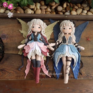 May include: Two handmade crochet fairy dolls with blonde hair and detailed wings. One doll has pink, white, and grey accents, while the other has blue and grey. Both dolls are on a wooden surface.
