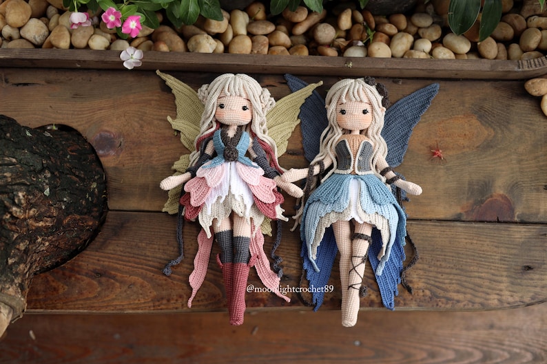 May include: Two handmade crochet fairy dolls with blonde hair and detailed wings. One doll has pink, white, and grey accents, while the other has blue and grey. Both dolls are on a wooden surface.
