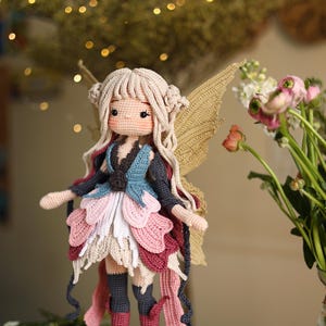 Peut inclure: Une poup&eacute;e f&eacute;e au crochet faite &agrave; la main, avec une peau claire, des cheveux blonds et de grandes ailes beiges. La poup&eacute;e porte une robe superpos&eacute;e dans les tons de rose, blanc et bleu, avec des bottes assorties. La poup&eacute;e est devant un fond flou avec des fleurs.