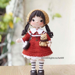 May include: Crocheted doll with brown hair, wearing a red dress, white socks with black stripes, and brown shoes. The doll is holding a brown crocheted teddy bear. The doll is standing on a wooden surface. The text "@moonlightcrochet89" is visible in the bottom right corner of the image.