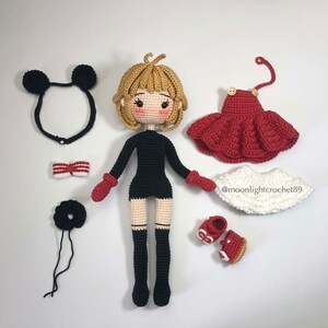 K&ouml;nnte beinhalten: Eine geh&auml;kelte Puppe mit blonden Haaren, die ein schwarzes Kleid, schwarze Stiefel und rote F&auml;ustlinge tr&auml;gt. Die Puppe ist von verschiedenen Accessoires umgeben, darunter eine rot-wei&szlig;e Schleife, ein schwarzes Stirnband mit Mickey-Mouse-Ohren, ein schwarzes Haargummi, ein rotes Kleid, ein wei&szlig;er Rock und rote Schuhe.