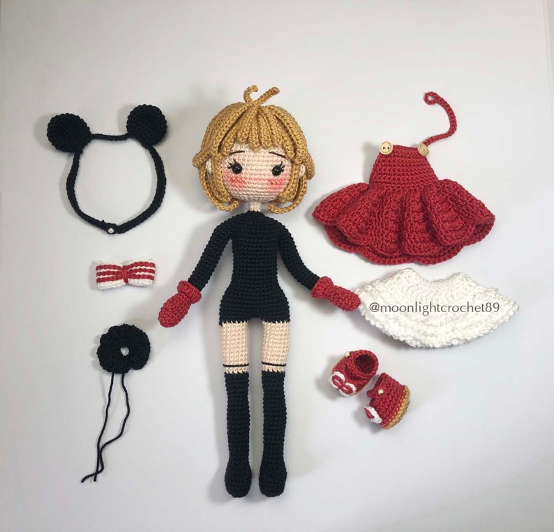 K&ouml;nnte beinhalten: Eine geh&auml;kelte Puppe mit blonden Haaren, die ein schwarzes Kleid, schwarze Stiefel und rote F&auml;ustlinge tr&auml;gt. Die Puppe ist von verschiedenen Accessoires umgeben, darunter eine rot-wei&szlig;e Schleife, ein schwarzes Stirnband mit Mickey-Mouse-Ohren, ein schwarzes Haargummi, ein rotes Kleid, ein wei&szlig;er Rock und rote Schuhe.