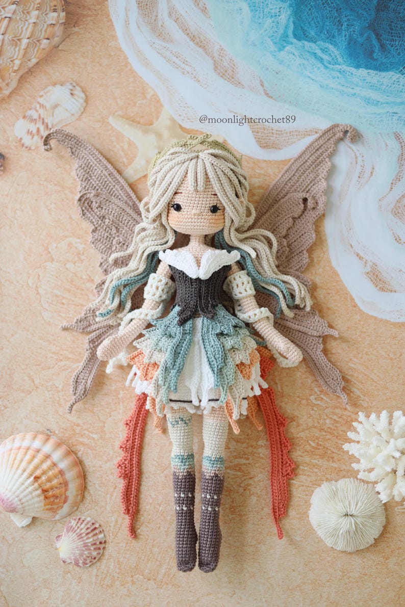 Puede incluir: Una mu&ntilde;eca de ganchillo con largo cabello ondulado color crema, grandes ojos oscuros y grandes alas marrones. La mu&ntilde;eca viste un corpi&ntilde;o marr&oacute;n oscuro, una falda a capas en tonos verde azulado, blanco y naranja, y botas altas marrones.