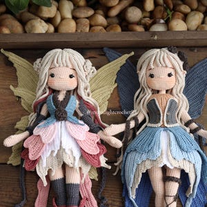 May include: Two handmade crochet fairy dolls with blonde hair, detailed dresses, and large wings. One doll wears a blue, pink, and white dress with green wings. The other doll has a blue dress and blue wings. The dolls are displayed on a wooden surface.