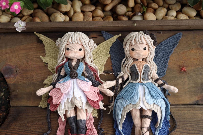 May include: Two handmade crochet fairy dolls with blonde hair, detailed dresses, and large wings. One doll wears a blue, pink, and white dress with green wings. The other doll has a blue dress and blue wings. The dolls are displayed on a wooden surface.