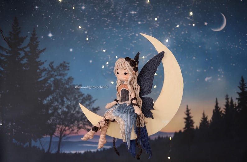 May include: A handcrafted crochet fairy doll with long blonde hair, a black floral headpiece, and blue wings sits on a crescent moon. The doll wears a blue and tan dress. The background is a night sky with stars and trees.