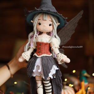 May include: A handmade crochet doll dressed as a fairy, wearing a gray witch hat with a teal brim, a rust-colored corset, and a gray skirt. The doll has gray wings and long, light purple hair. The doll is held by a person. The text @moonlightcrochet89 is visible.
