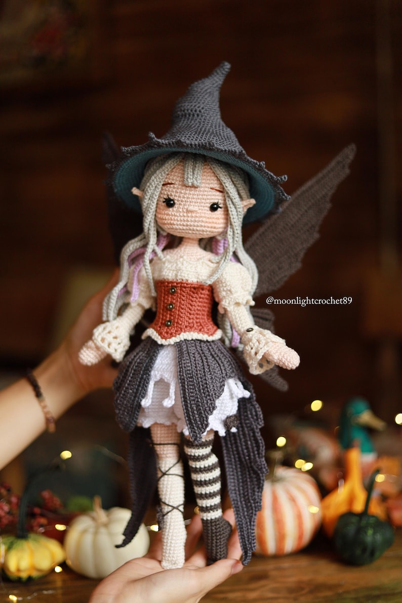 May include: A handmade crochet doll dressed as a fairy, wearing a gray witch hat with a teal brim, a rust-colored corset, and a gray skirt. The doll has gray wings and long, light purple hair. The doll is held by a person. The text @moonlightcrochet89 is visible.