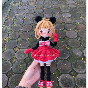 K&ouml;nnte beinhalten: Geh&auml;kelte Puppe mit schwarzer und roter Kleidung, schwarzen Haaren und einer roten Schleife. Die Puppe hat einen Mickey-Mouse-artigen Kopf mit gro&szlig;en schwarzen Ohren. Die Puppe h&auml;lt einen roten F&auml;ustling in ihrer rechten Hand. Die Puppe steht auf einer grauen und roten Ziegelstein-Oberfl&auml;che. Der Text "@moonlightcrochet89" ist auf dem Kleid der Puppe sichtbar.