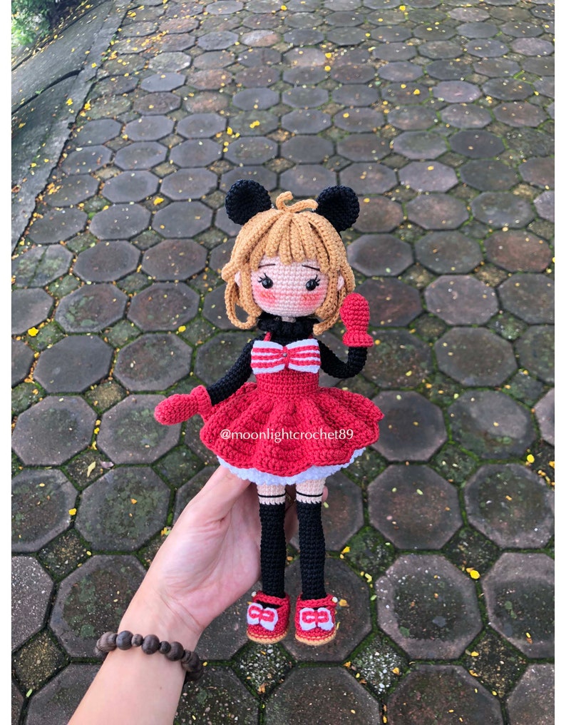K&ouml;nnte beinhalten: Geh&auml;kelte Puppe mit schwarzer und roter Kleidung, schwarzen Haaren und einer roten Schleife. Die Puppe hat einen Mickey-Mouse-artigen Kopf mit gro&szlig;en schwarzen Ohren. Die Puppe h&auml;lt einen roten F&auml;ustling in ihrer rechten Hand. Die Puppe steht auf einer grauen und roten Ziegelstein-Oberfl&auml;che. Der Text "@moonlightcrochet89" ist auf dem Kleid der Puppe sichtbar.