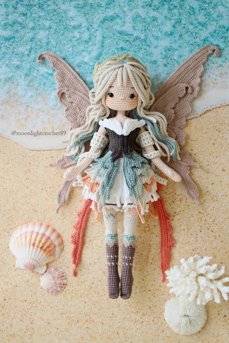 Puede incluir: Una mu&ntilde;eca de ganchillo con cabello largo y ondulado, alas grandes y un atuendo en capas en tonos beige, blanco, azul verdoso y coral. La mu&ntilde;eca tiene un corpi&ntilde;o oscuro, pu&ntilde;os en los brazos y botas altas. El texto @moonlightcrochet89 es visible a la izquierda.